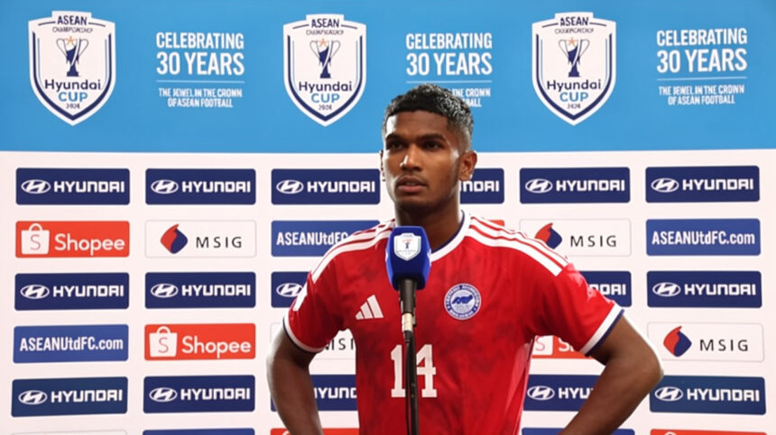 Soccer player in a red jersey with number 14 speaks at a press conference, blue backdrop showing ASEAN Cup logos and sponsors behind him.