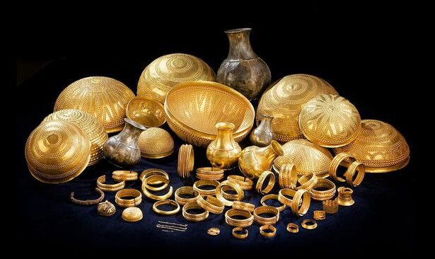 A display of gold jewelry and decorative bowls arranged on a dark surface, including bracelets, rings, bangles, and vases.
