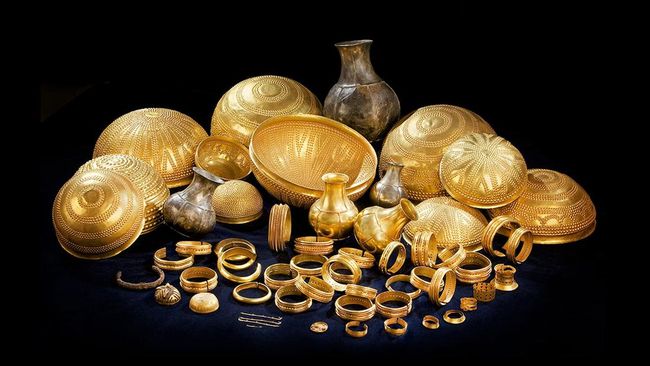A display of gold jewelry and decorative bowls arranged on a dark surface, including bracelets, rings, bangles, and vases.