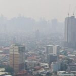 Hazy city skyline with a tall, under-construction skyscraper and cranes rising above the other buildings