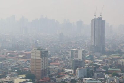 Hazy city skyline with a tall, under-construction skyscraper and cranes rising above the other buildings