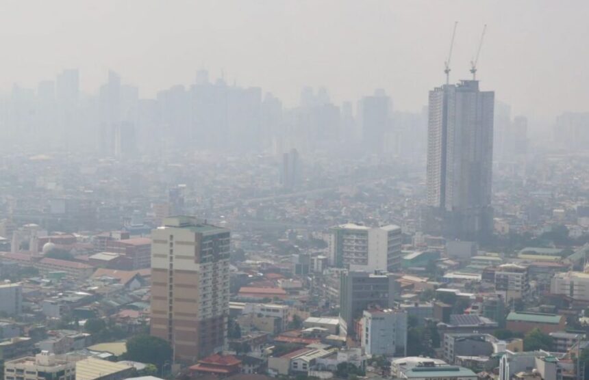 Hazy city skyline with a tall, under-construction skyscraper and cranes rising above the other buildings