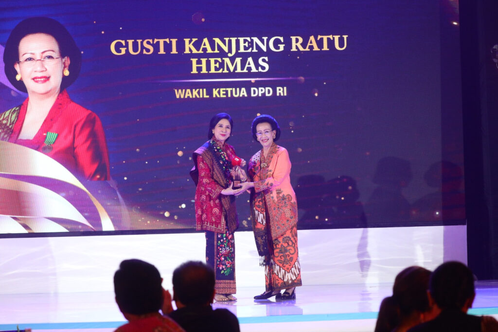 Two women in colorful batik on a lit stage exchange an award, with a large backdrop displaying a portrait and Indonesian text behind them.