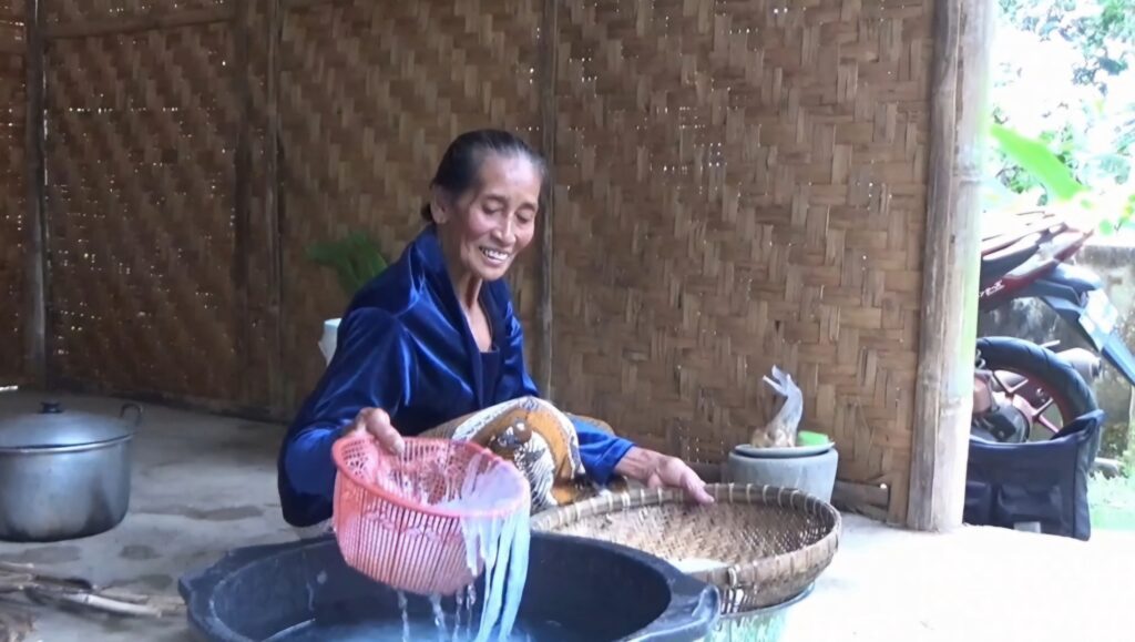 hghg - Mabur.co Elderly woman in a blue jacket weaving a large wicker basket outdoors beside a woven bamboo wall.