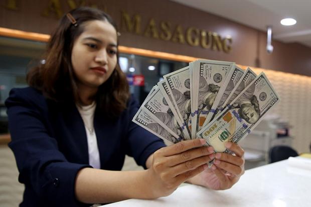 Woman in a blazer at a bank counter fanning out multiple US hundred-dollar bills in her hands.