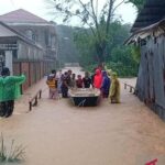 Group of villagers guiding a small boat through a flood that covers a street between houses, wearing colorful raincoats and ponchos.