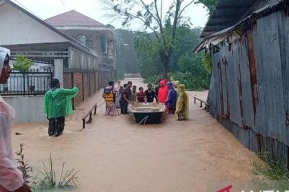 Group of villagers guiding a small boat through a flood that covers a street between houses, wearing colorful raincoats and ponchos.