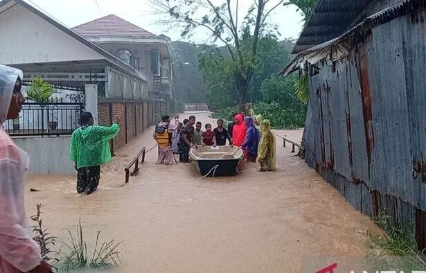 Group of villagers guiding a small boat through a flood that covers a street between houses, wearing colorful raincoats and ponchos.