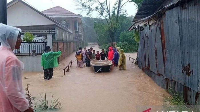 Group of villagers guiding a small boat through a flood that covers a street between houses, wearing colorful raincoats and ponchos.