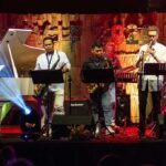 Live band performing on stage: pianist at left, saxophonists in center, trumpet player at right, with colorful stage lighting.