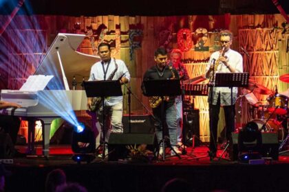 Live band performing on stage: pianist at left, saxophonists in center, trumpet player at right, with colorful stage lighting.