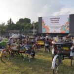 Outdoor festival scene with a large stage screen reading 'Kotabaru Heritage Festival' and many colorful rickshaws and chairs on a grassy field.