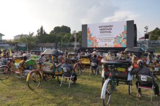 Outdoor festival scene with a large stage screen reading 'Kotabaru Heritage Festival' and many colorful rickshaws and chairs on a grassy field.