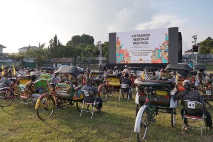Outdoor festival scene with a large stage screen reading 'Kotabaru Heritage Festival' and many colorful rickshaws and chairs on a grassy field.