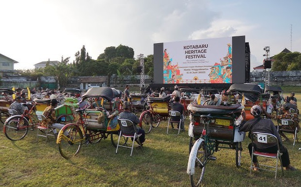 Outdoor festival scene with a large stage screen reading 'Kotabaru Heritage Festival' and many colorful rickshaws and chairs on a grassy field.