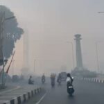 A hazy urban street scene with several motor scooters riding toward the camera and a few pedestrians on the sidewalk; a tall column-like monument is visible in the distance.
