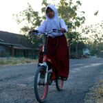 A young girl wearing a white hijab and red skirt rides a bicycle on a rural road with houses and trees in the background.