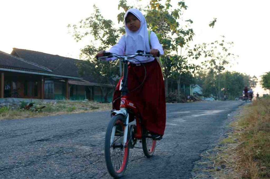 A young girl wearing a white hijab and red skirt rides a bicycle on a rural road with houses and trees in the background.