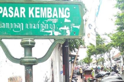 Green street sign reading 'Jl. Pasar Kembang' on a weathered post, with a busy market street and trees in the background.
