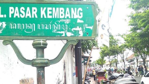 Green street sign reading 'Jl. Pasar Kembang' on a weathered post, with a busy market street and trees in the background.