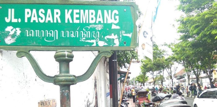Green street sign reading 'Jl. Pasar Kembang' on a weathered post, with a busy market street and trees in the background.