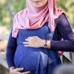 Pregnant woman wearing a pink hijab and blue denim maternity dress stands by a wooden fence, cradling her baby bump outdoors.