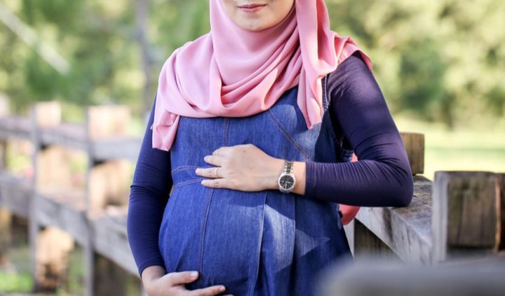 Pregnant woman wearing a pink hijab and blue denim maternity dress stands by a wooden fence, cradling her baby bump outdoors.