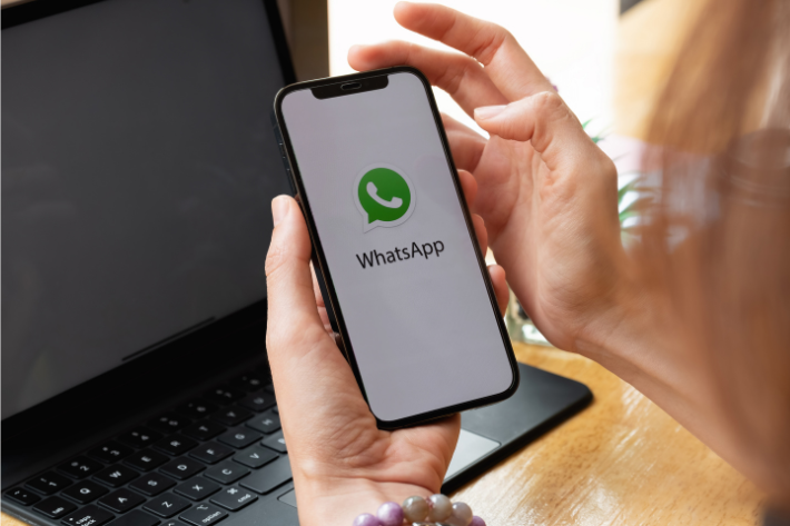Hand holding a smartphone displaying the WhatsApp app screen beside a laptop.