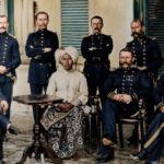 Group portrait of nine officers in dark military uniforms with gold detailing, gathered around a central seated elderly woman in a white headwrap and patterned dress, outdoors in a courtyard.