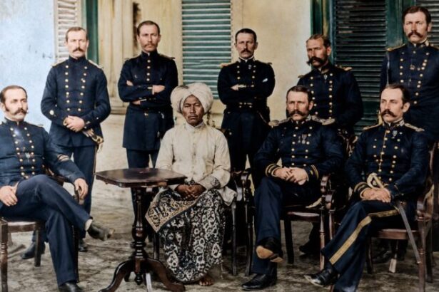 Group portrait of nine officers in dark military uniforms with gold detailing, gathered around a central seated elderly woman in a white headwrap and patterned dress, outdoors in a courtyard.