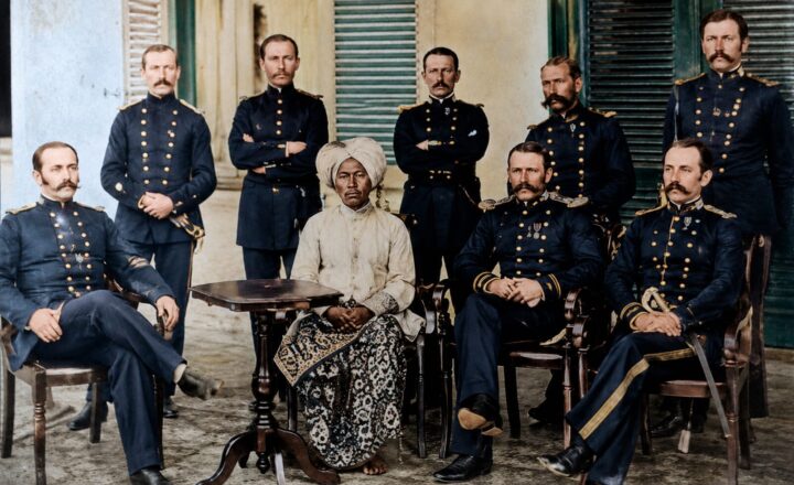 Group portrait of nine officers in dark military uniforms with gold detailing, gathered around a central seated elderly woman in a white headwrap and patterned dress, outdoors in a courtyard.