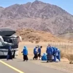 Bus with front-end crash on desert highway; people in blue outfits gather nearby, mountain backdrop behind a fenced area.