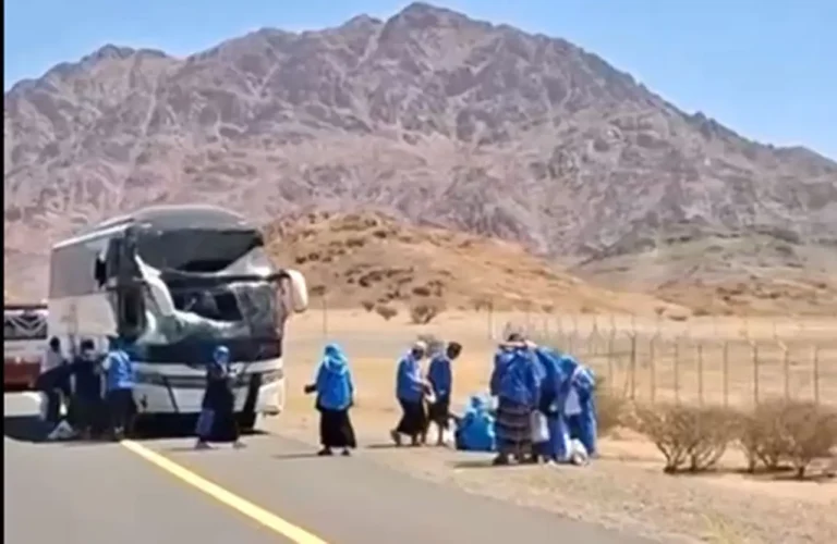 Bus with front-end crash on desert highway; people in blue outfits gather nearby, mountain backdrop behind a fenced area.