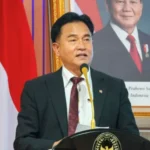 A man in a dark suit speaking at a podium with a gold emblem, Indonesian flag behind him, and a framed portrait in the background.