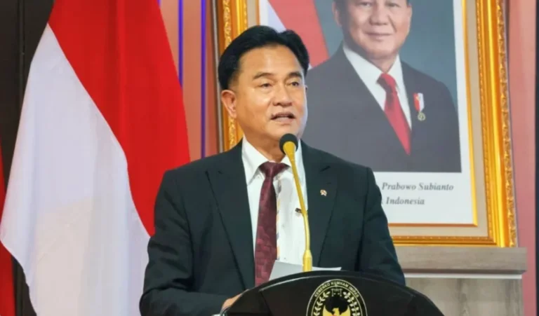 A man in a dark suit speaking at a podium with a gold emblem, Indonesian flag behind him, and a framed portrait in the background.