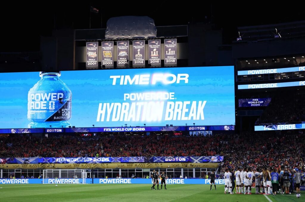 Large stadium jumbotron reads 'TIME FOR HYDRATION BREAK' with Powerade ad; soccer match underway behind the field banners and fans in stands.