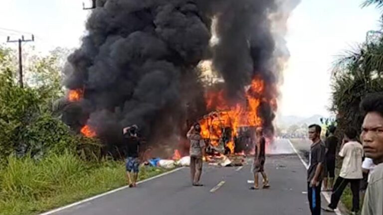 Bus on fire in the middle of a road, large black smoke plumes rising as bystanders watch nearby