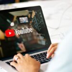 Person typing on a laptop displaying the YouTube logo and a video camera, suggesting video creation or uploading to YouTube