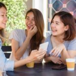 Three friends sit at a round outdoor table, smiling and looking at a smartphone together, with coffee cups nearby.