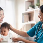 Pediatric nurse in teal scrubs smiling at a mother and her baby during a check-up in a clinic setting.