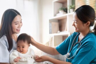 Pediatric nurse in teal scrubs smiling at a mother and her baby during a check-up in a clinic setting.