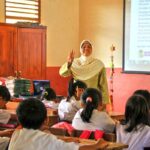 Teacher in a hijab stands at the front of a classroom, making a peace sign as students listen attentively.