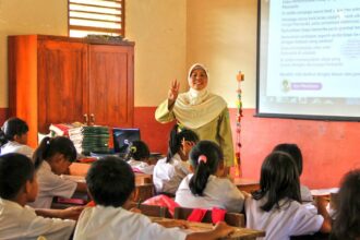 Teacher in a hijab stands at the front of a classroom, making a peace sign as students listen attentively.