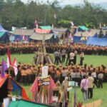 Large group of scouts forming a circle around a central wooden bonfire on a grassy field, with colorful tents and flags in the background.