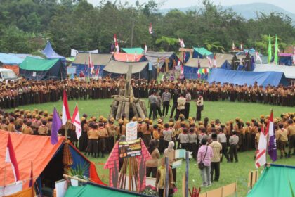 Large group of scouts forming a circle around a central wooden bonfire on a grassy field, with colorful tents and flags in the background.
