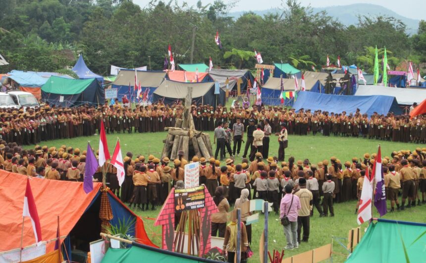 Large group of scouts forming a circle around a central wooden bonfire on a grassy field, with colorful tents and flags in the background.
