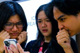 Three young people gathered around a smartphone, frowning and looking concerned.
