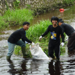 Group of volunteers wading in a river, collecting litter into a white bag by the grassy bank.
