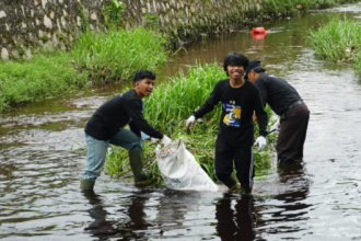 Group of volunteers wading in a river, collecting litter into a white bag by the grassy bank.