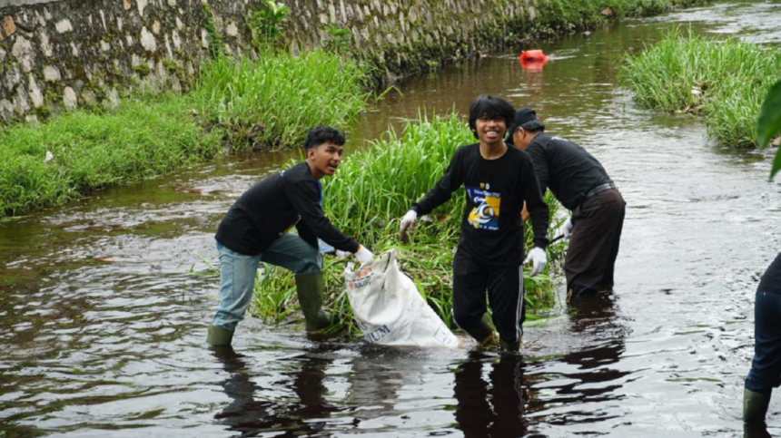Group of volunteers wading in a river, collecting litter into a white bag by the grassy bank.